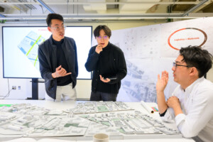 Two people stand in front of a table on which sits a model of an urban landscape. A third person sits at the table. City plans and maps are pinned to a wall behind them and displayed on a screen.