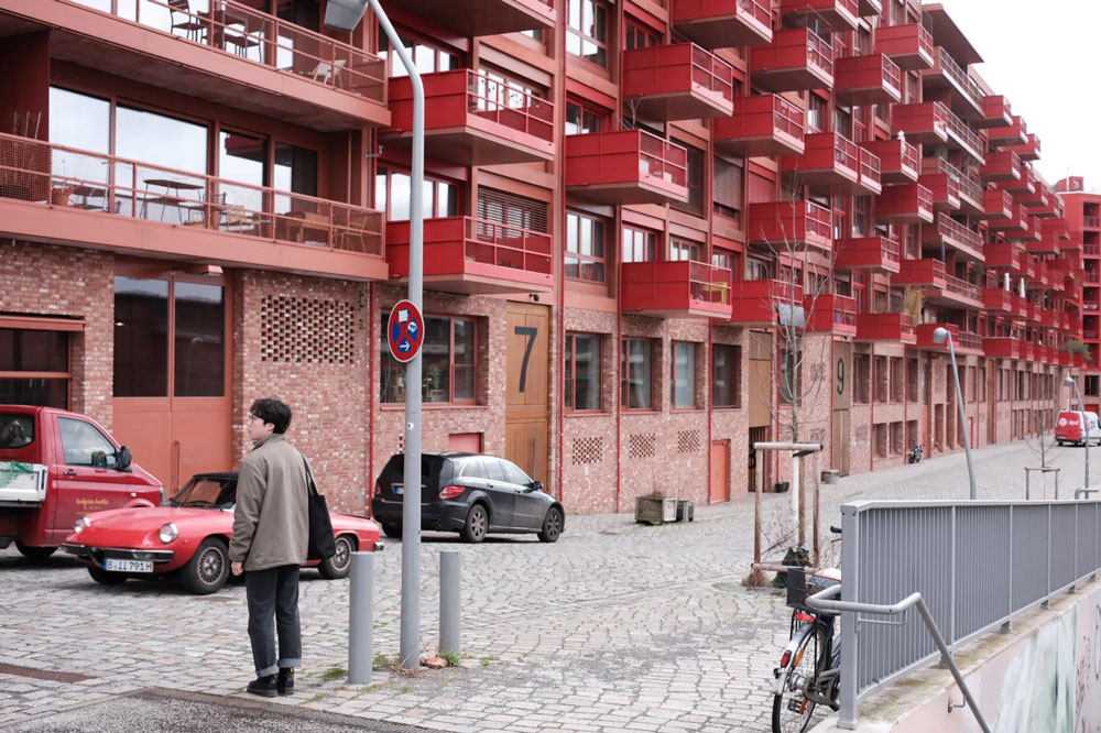 An individual stands in front of an apartment building that appears to stretch for a city block and is about five or six stories high. Many Red balconies protrude from the building.