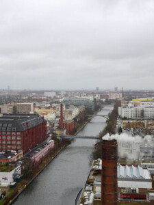 A photograph taken from a high position shows a canal in an urban area with a mix of large buildings including structures made of brick and others made of glass and steel. Several buildings have smokestacks.
