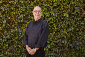 A portrait of Peter Walker who is dressed in dark clothes and stands in front of a wall of green plants.