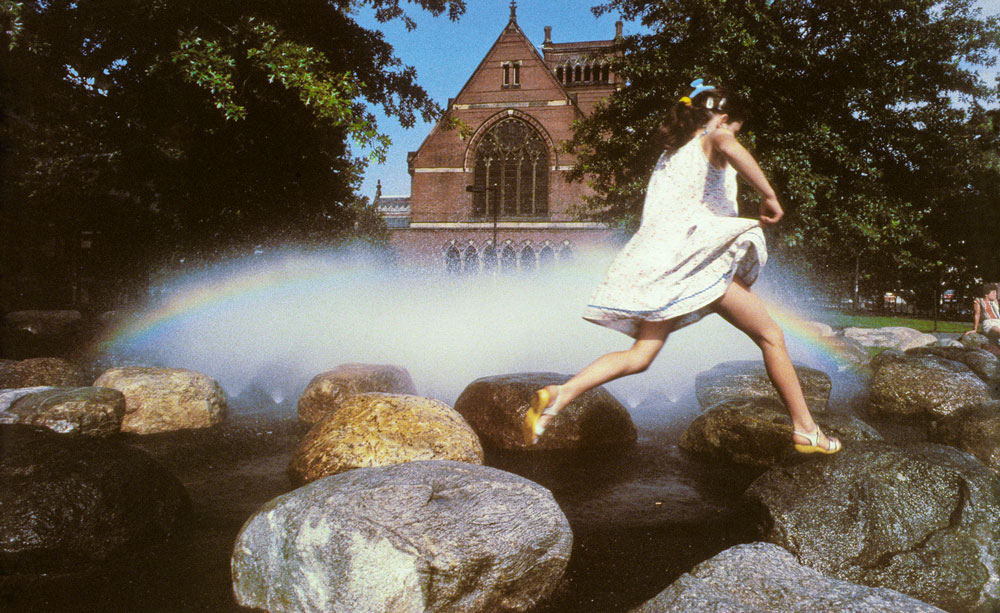 A vintage photograph shows a girl in a white dress jumping on rocks. A fountain sprays mist on the rocks.