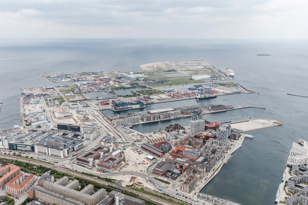 An aerial view of a large waterfront area of Copenhagen. The area includes dense neighborhoods of large buildings, industrial docks with ships next to them, and open spaces near the sea.