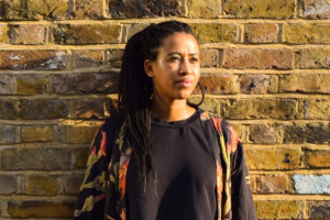 A portrait of Thandi Loewenson who stands in front of a brick wall.