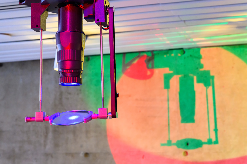 A color image of a projector, the lens of which is pointed at a rotating mirror. Colorful lights are visible on the wall in the background.