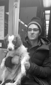 Will Conroy holding a dog while sitting on the subway