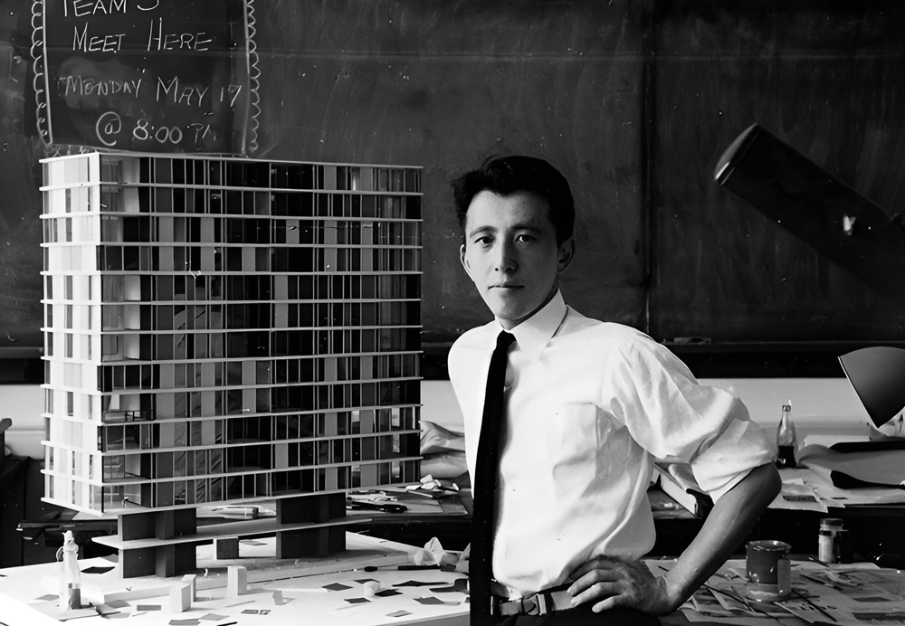 A black-and-white portrait of Fumihiko Maki posing in front of an architectural model of a high-rise building. He wears a white shirt and a dark tie.