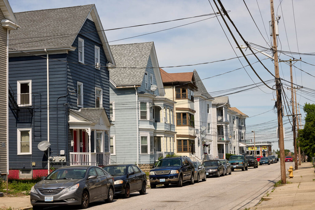 A photo of a street with triple decker houses on the left side and with cars parked in front of the houses.