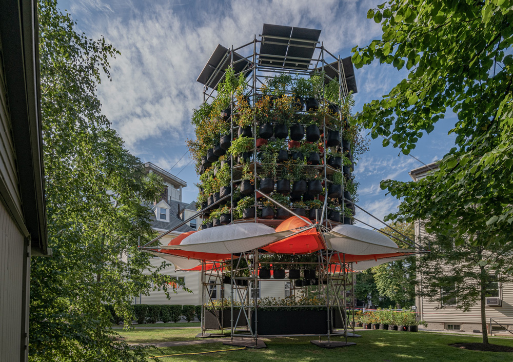 A photograph of Belinda Tato's Polinature project. A four-story cylindrical structure of scaffolding supports tiers of plants in black grow bags. Orange and white inflated bubbles ring the lower level.