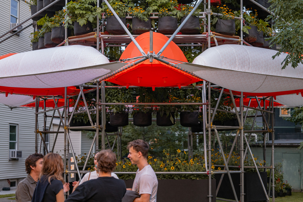 A photograph of Belinda Tato's Polinature project. A four-story cylindrical structure of scaffolding supports tiers of plants in black grow bags. Orange and white inflated bubbles ring the lower level. People stand around the structure and chat.