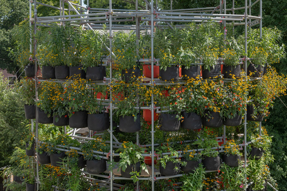 A photograph of Belinda Tato's Polinature project. A four-story cylindrical structure of scaffolding supports tiers of plants in black and orange grow bags.