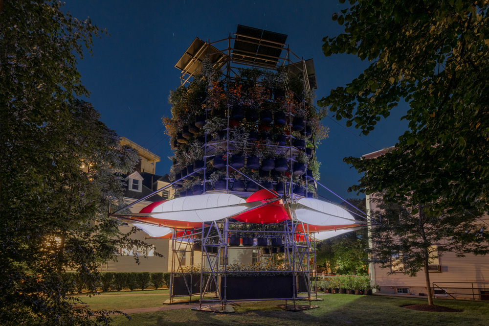 A photograph of Belinda Tato's Polinature project. A four-story cylindrical structure of scaffolding supports tiers of plants in black grow bags. Orange and white inflated bubbles ring the lower level.