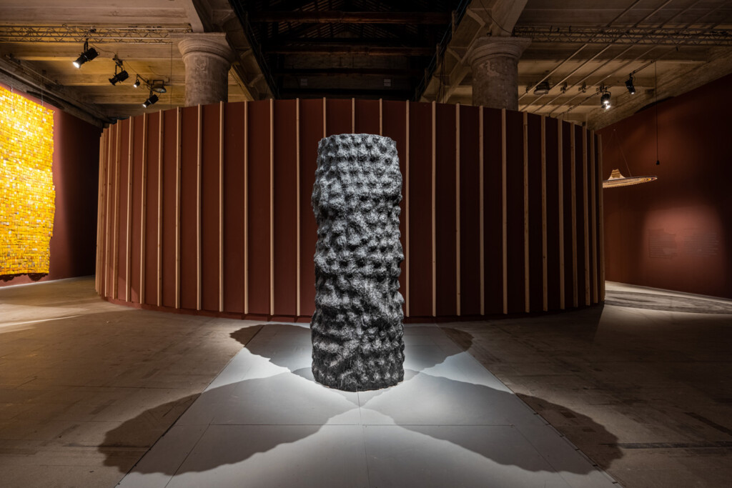 A view of a black column by Germane Barnes standing in an exhibition space in the Venice Biennale. The column has a knotty, irregular surface.