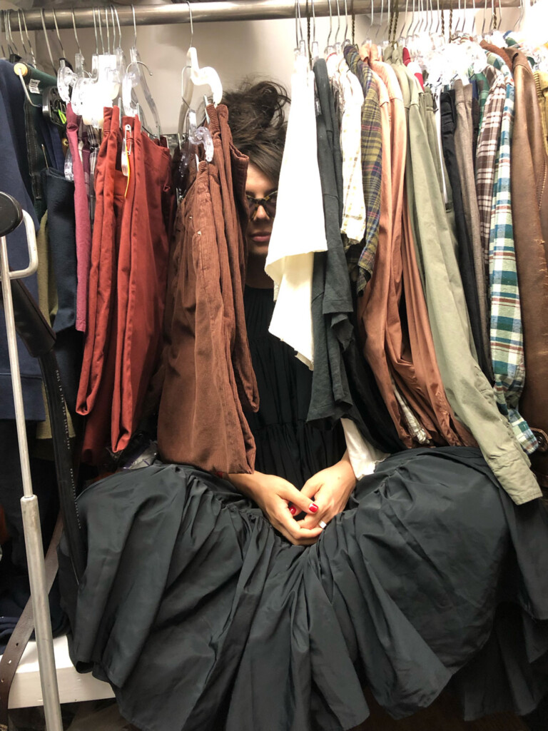 Stacey Berman appears wearing a black dress sitting down below a clothing rack. She is partially obscured by hanging clothing.