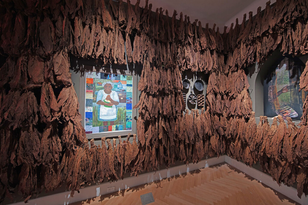 A view of a room with walls lined in tobacco leaves. There are paintings hanging in openings on the wall and a display of cast-iron cookware.