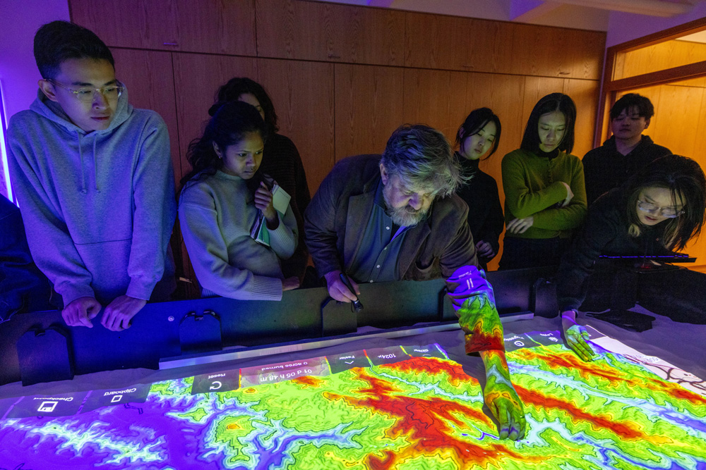 Man and students look at colorful map on sand table